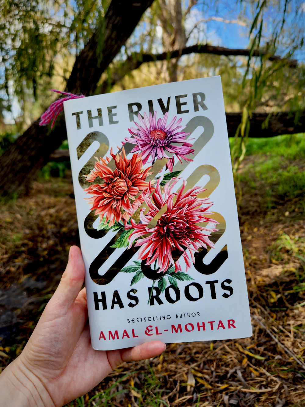 The book The River Has Roots by Amal El-Mohtar, which has three pink flowers on the cover, held up before a willow tree leaning over a creek.