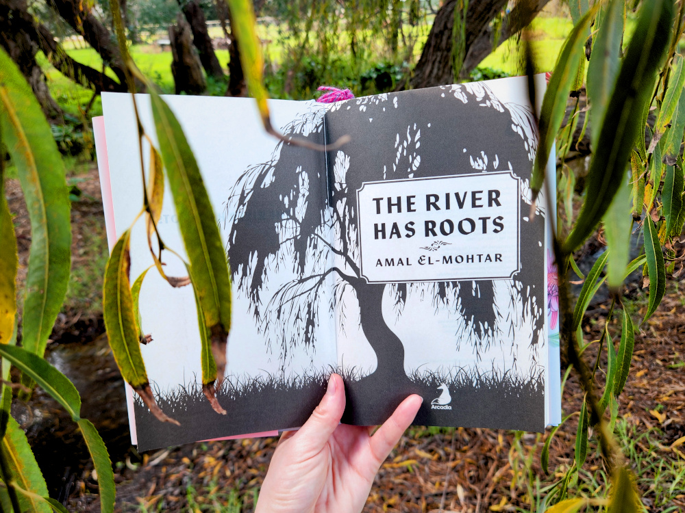 The title page of The River Has Roots by Amal El-Mohtar, which features a black and white illustration of a willow tree, held up between weeping willow leaves.