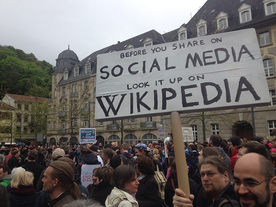 A crowd of people in a city square. A man is holding a sign that reads "Before you share on social media, look it up on Wikipedia."