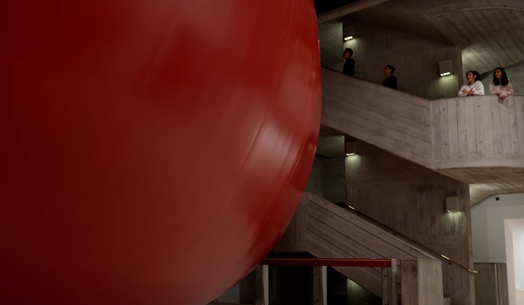 A high-angle view from an internal concrete stairway looking down at a massive, deep-red inflated structure that fills and obstructs a gallery entrance hall.