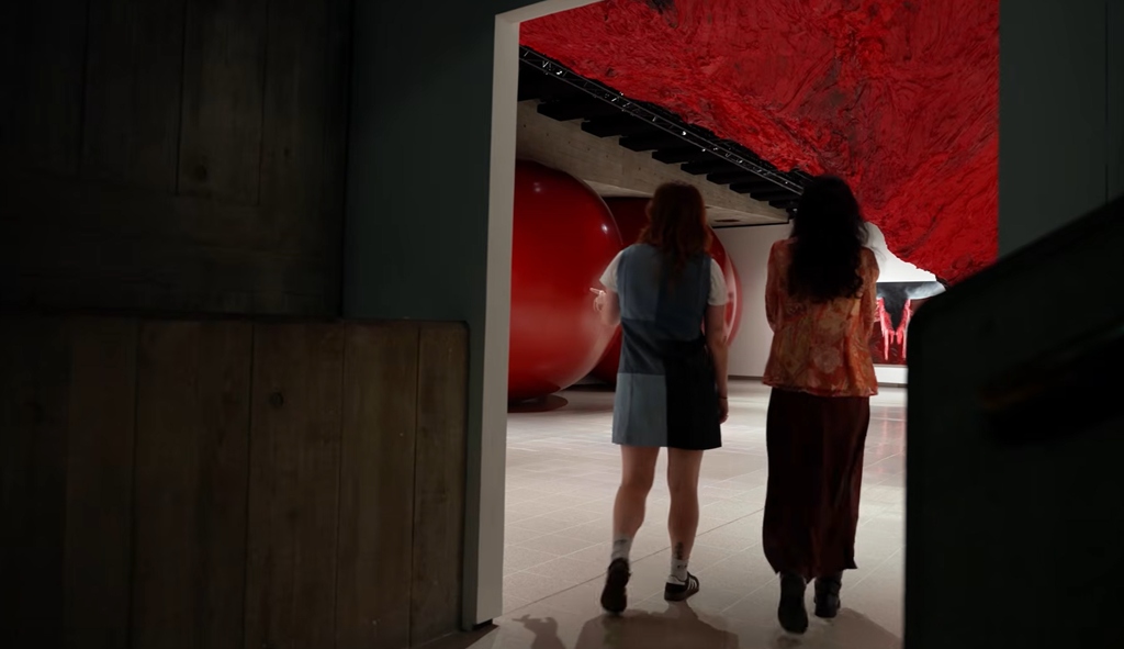 An eye-level view of two people standing beneath a gigantic red and black sculptural structure after passing through the entrance to the gallery space.