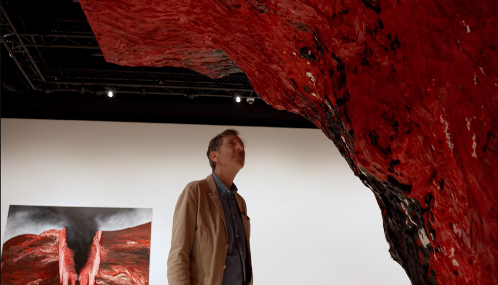 An eye-level view looking up at an enormous, upside-down red and black sculpture projecting downward from the ceiling.