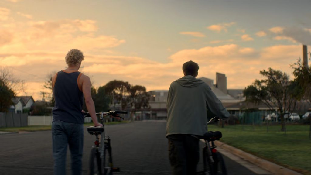 Two teenage boys walk their bicycles down a quiet suburban street at sunset, with houses, trees and open grassy verges around them beneath a glowing orange sky. Seen from behind, they move side by side through the otherwise empty street.