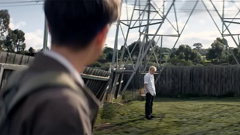 A young man in the foreground looks towards another young man standing alone in a grassy backyard, surrounded by weathered timber fencing and a large metal power structure.